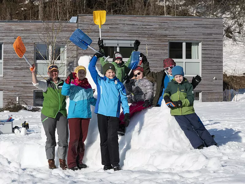Familie steht im Schnee mit Schneeschaufeln vor Holzhaus