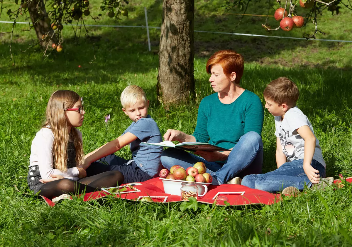 Guide und drei Kinder sitzen mit einem Buch und Äpfeln auf einem Tuch in der Wiese unter Apfelbäumen