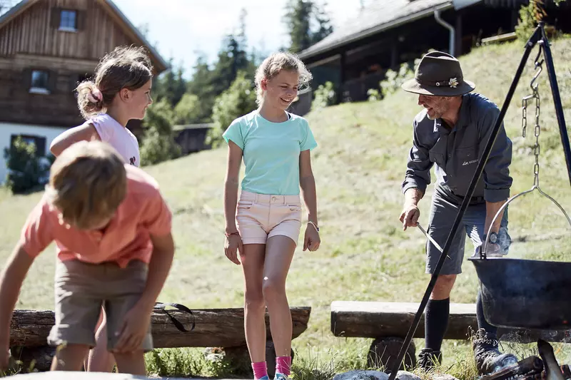 3 Kinder stehen mit einem Ranger bei Holzbänken an einem Kessel über einem Lagerfeuer